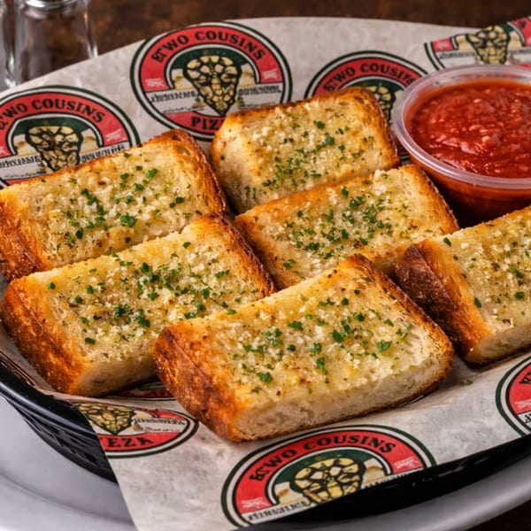 A basket of Two Cousins Pizza garlic bread with parsley and parmesan, served with a side of marinara sauce.